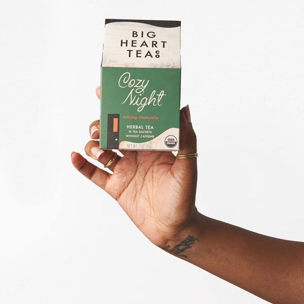 Hand holding the Cozy Night tea, blended with organic chamomile, organic spearmint, organic lemongrass. The box is green and made to look like a cottage in the woods on a snowy day. 